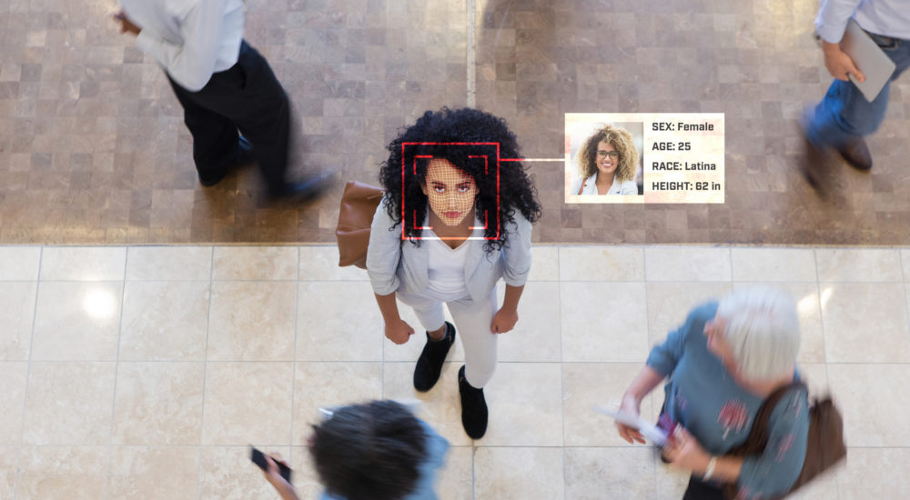A young Hispanic businesswoman looks up while in an office lobby with businesspeople all around her. A facial recognition scan reveal her personal data.